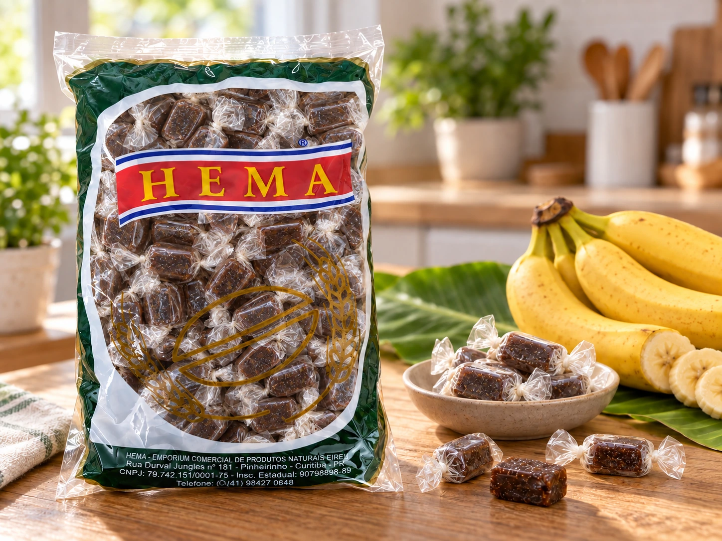 Pacote HEMA de bala de banana com moldura verde, faixa HEMA e desenho dourado na frente, balas em invólucros individuais visíveis pela embalagem, ao lado de tigela com balas e bananas frescas